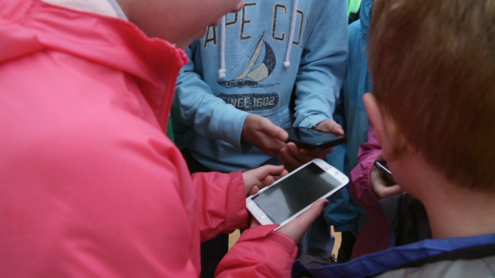 A group of children clustered together on a school field trip, working through a spy challenge on phones together