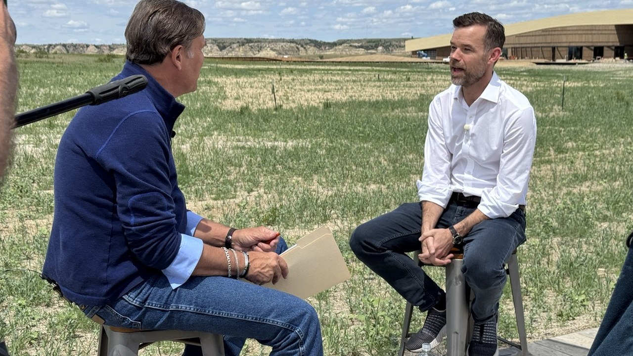 A network correspondent interviewing a Library executive on stools outdoors, the building visible behind them