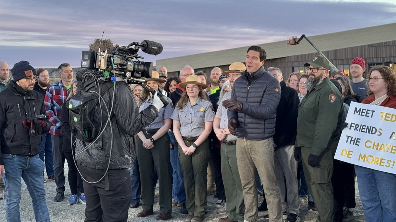 A network correspondent delivering a live segment at dawn, surrounded by National Park Service rangers, Library staff and Medora residents holding signs
