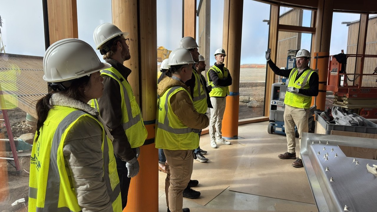 A hard-hat walkthrough of the unfinished Library interior, a group in high-visibility vests listening beside mass timber columns