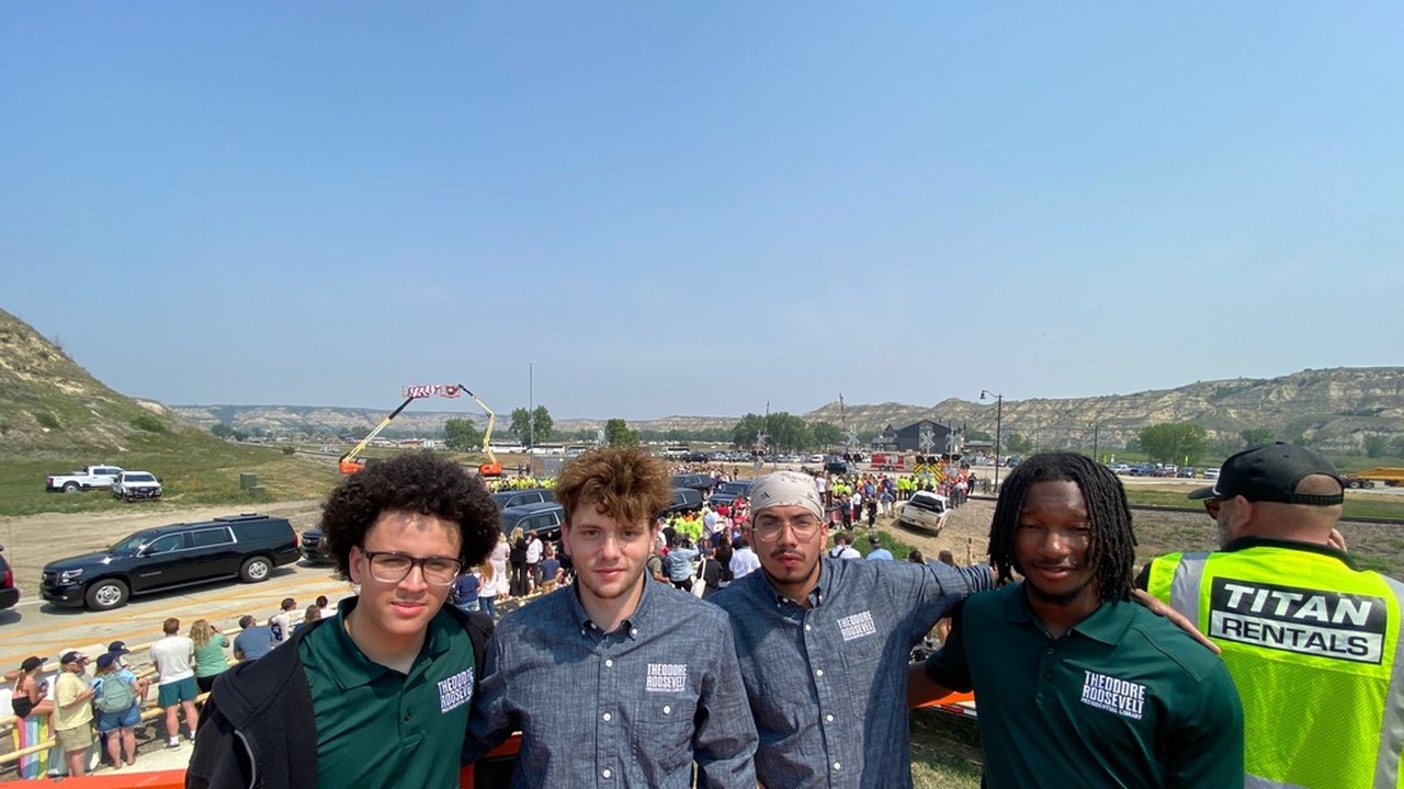 Four Theodore Roosevelt Presidential Library interns standing together on the media riser, the motorcade and crowd visible on the road below